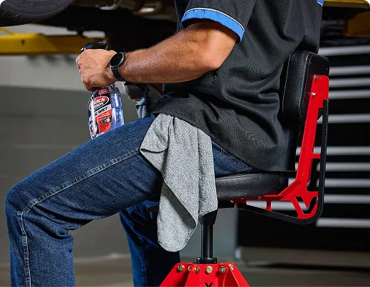 A mechanic sits on a red shop stool holding a spray bottle and a cloth.