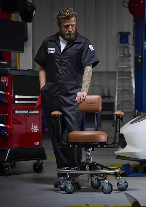 A bearded mechanic in a black jumpsuit stands next to a brown leather rolling stool in a garage.