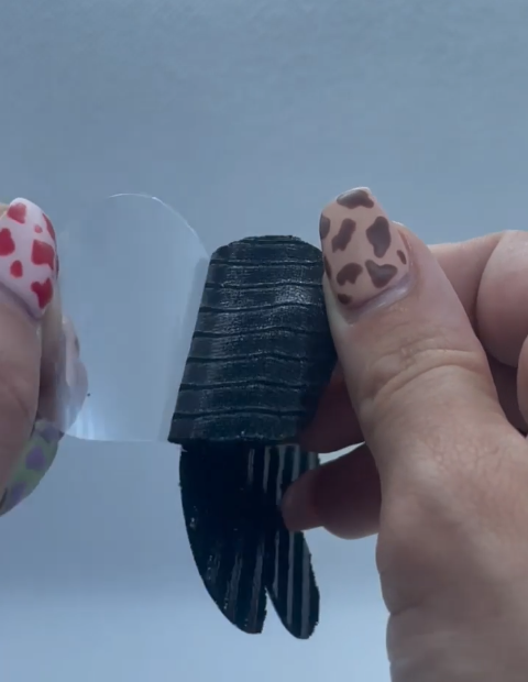 A person with decorated nails peels a clear film from a black, textured, and striped object.