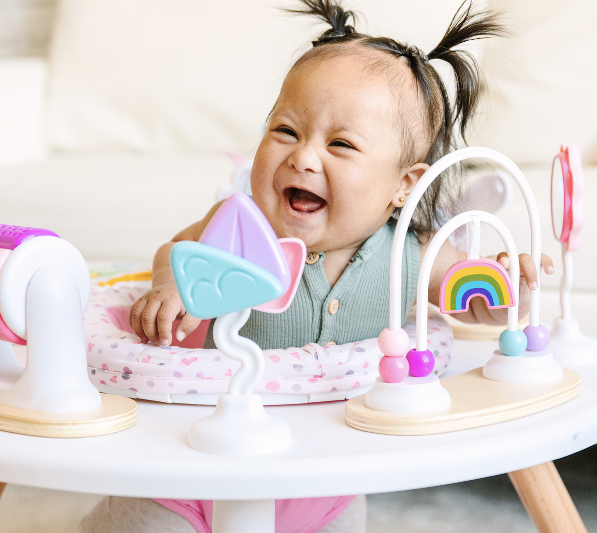 A laughing baby with pigtails sits in a colorful activity center.