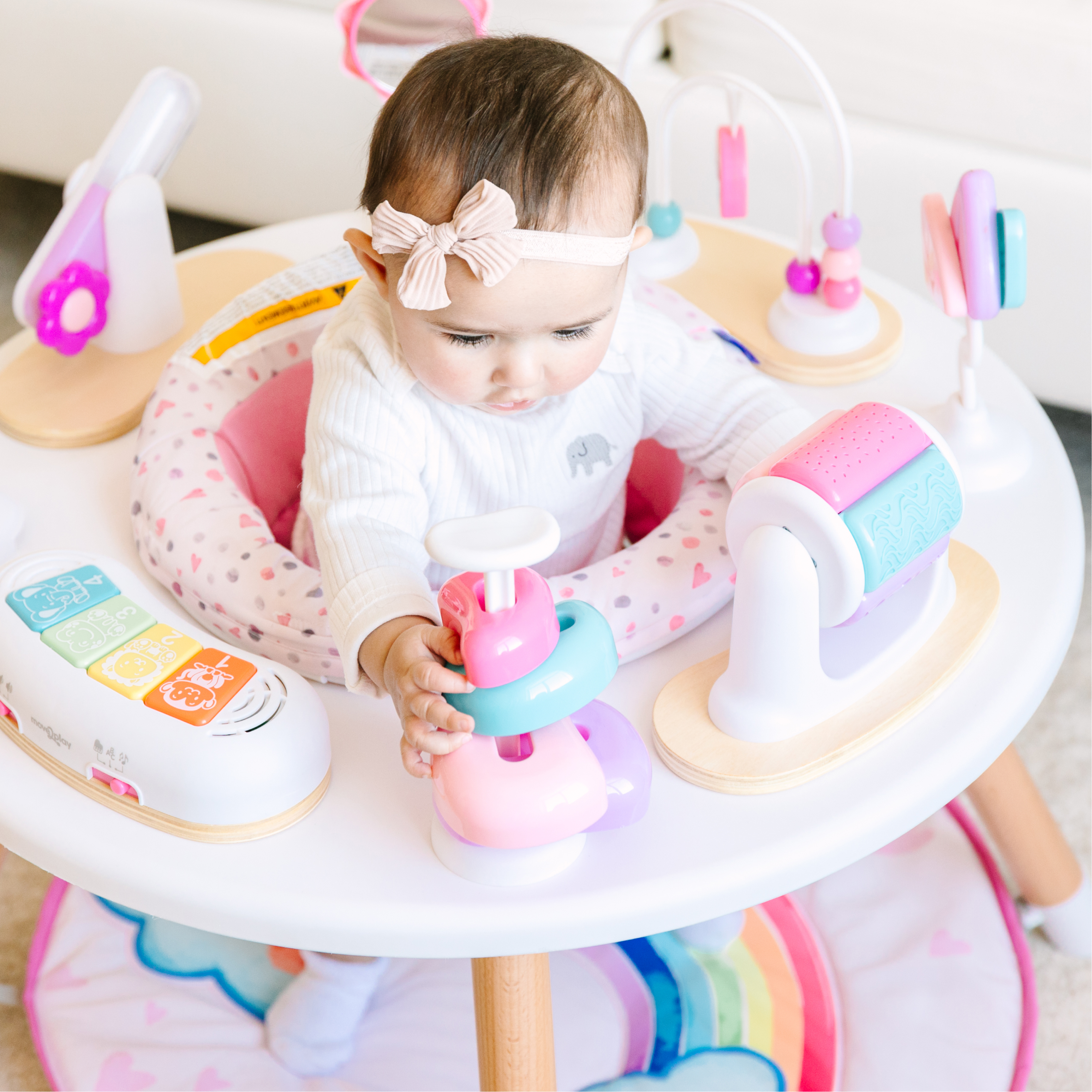 A baby girl with a pink bow sits in a white activity center playing with toys.