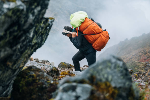 Hiker on a rocky mountain path carrying large backpacks in foggy conditions.
