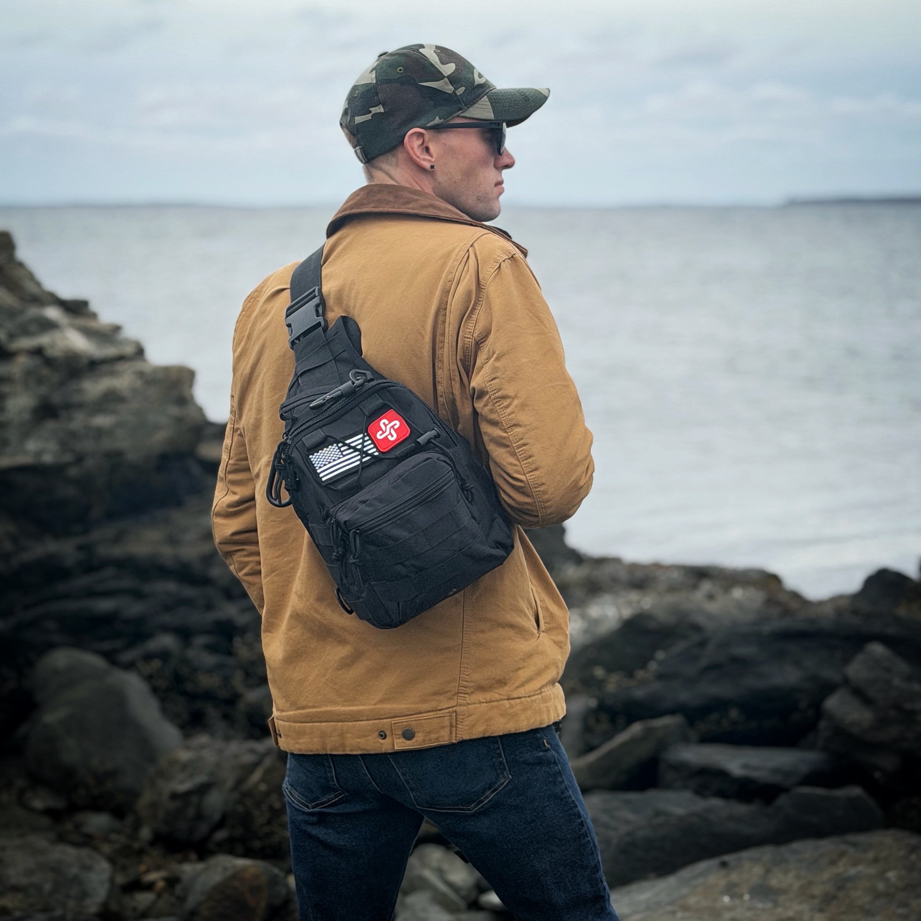 Person with a backpack stands by rocky waterfront wearing a cap and brown jacket.
