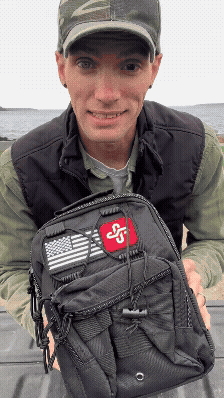 Person holding a black tactical backpack with patches, outdoors.