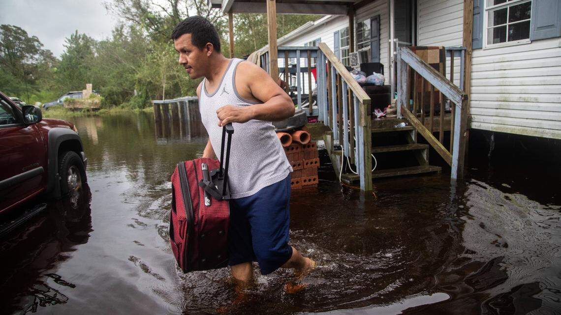Person carrying luggage through floodwater near a house and car.