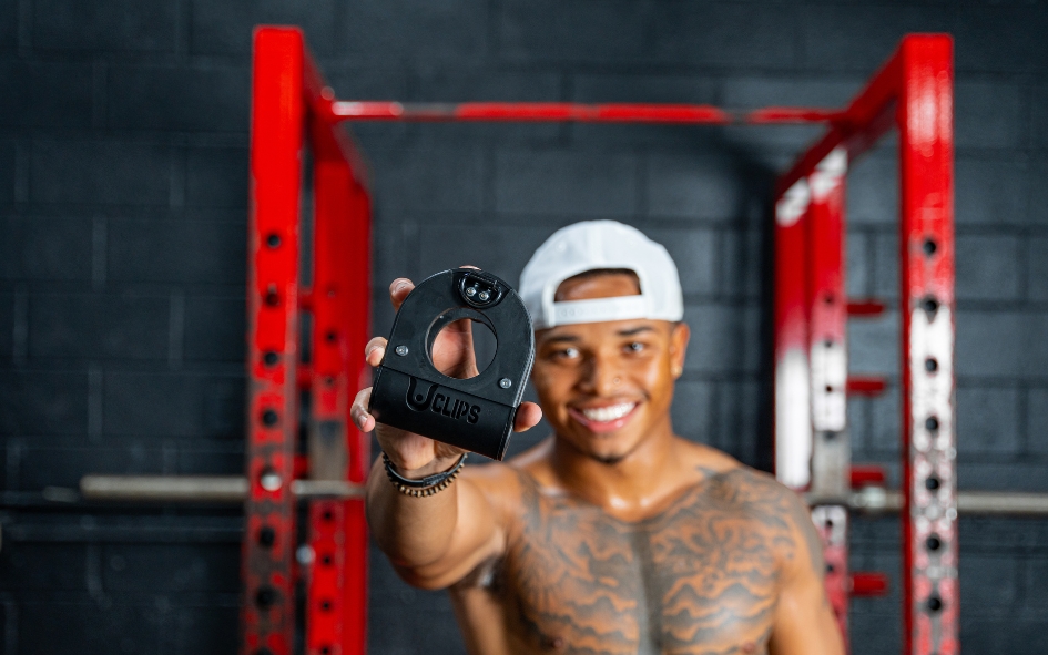 Man in gym holding a black clip, smiling, with red gym equipment in background.