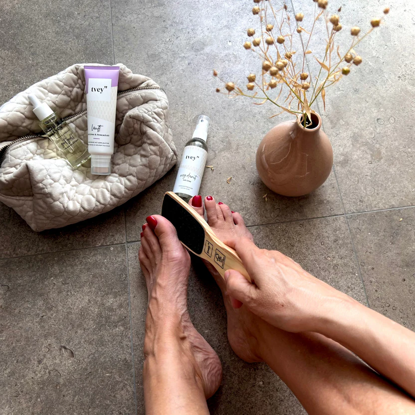 Person using a foot file, with skincare products and a vase on the floor.