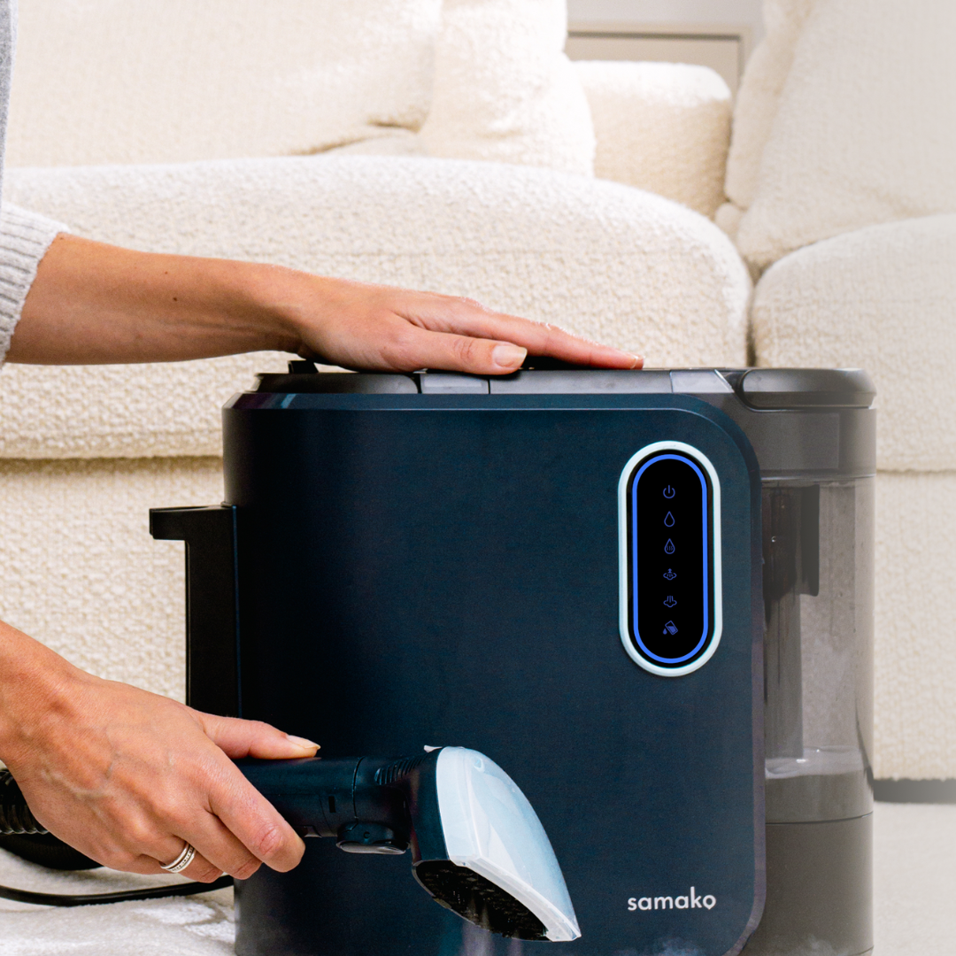 A person using a dark blue portable upholstery cleaner with a brush nozzle next to a cream-colored sofa.