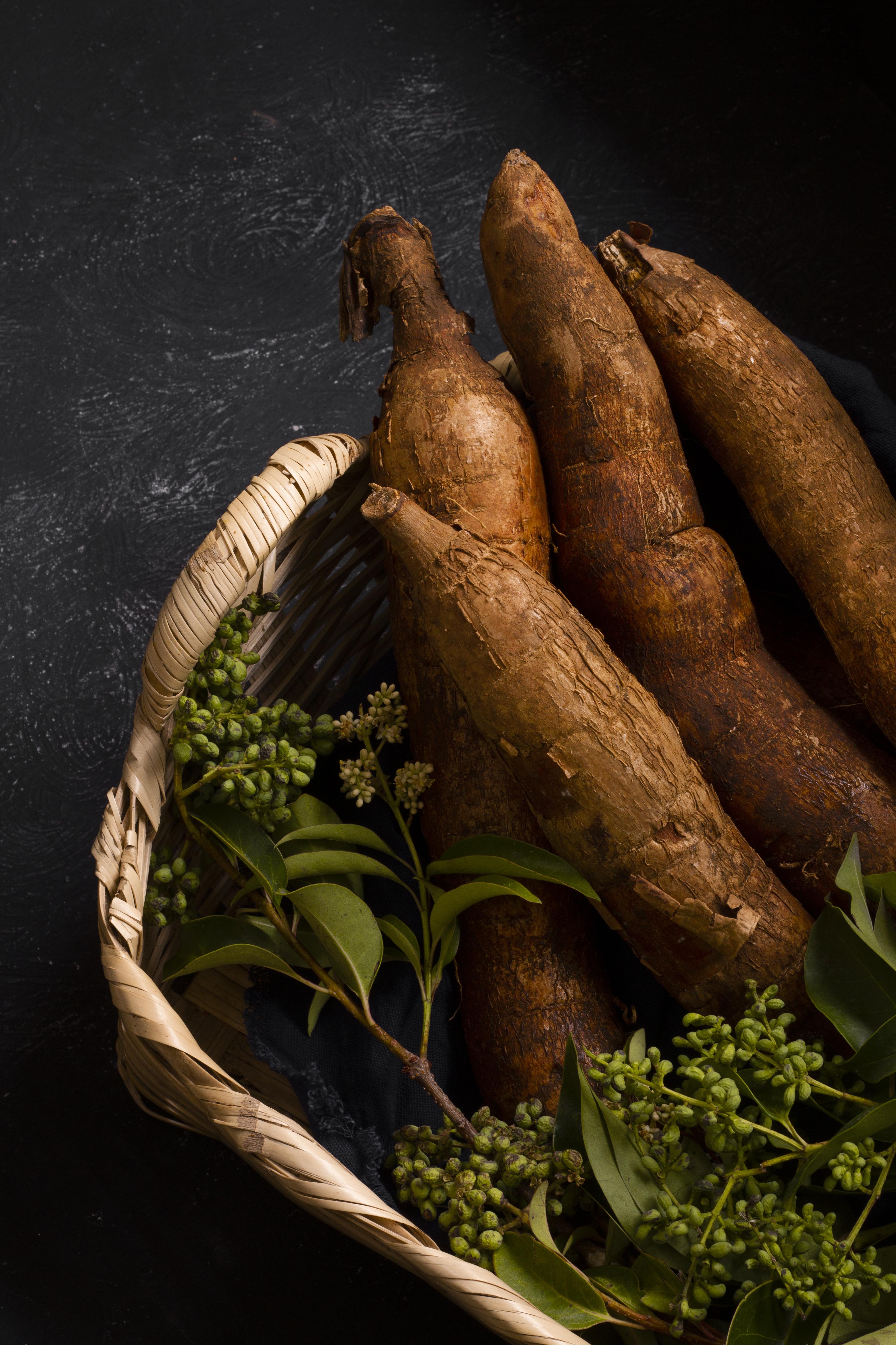 A top-down view of a woven basket filled with cassava roots and green sprigs on a dark surface.