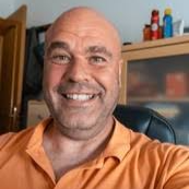 Smiling man in an orange shirt sitting indoors.