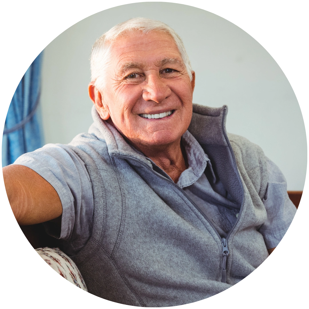 A smiling elderly man with white hair, wearing a grey zip-up fleece, sitting indoors.