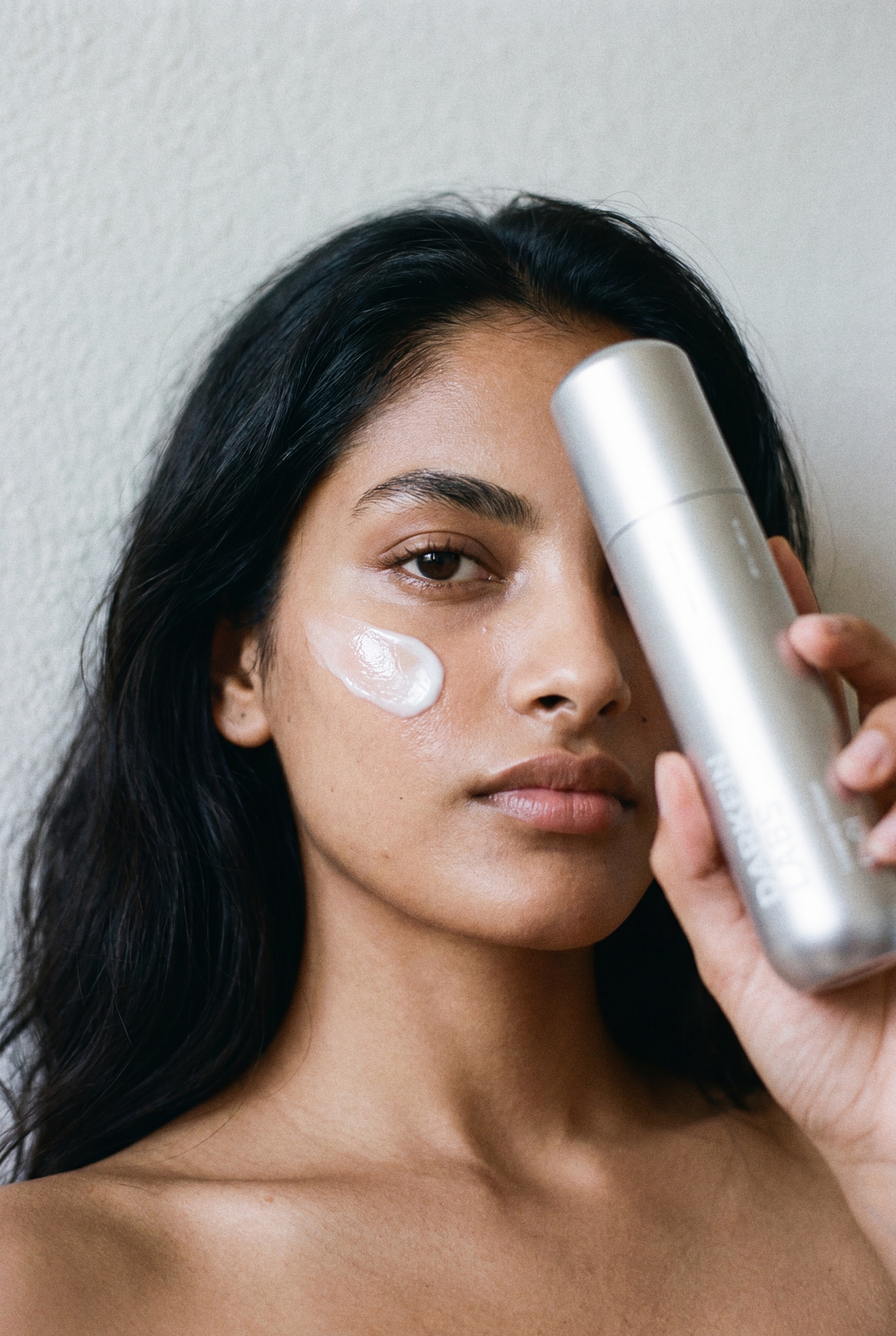 A woman with a dab of white cream on her cheek holds a silver bottle partially over her face.
