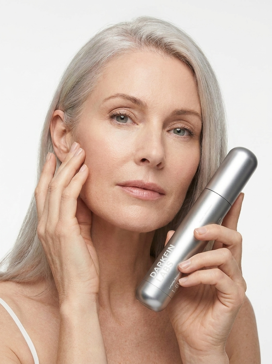 A woman with grey hair holds a silver skincare bottle while touching her face.