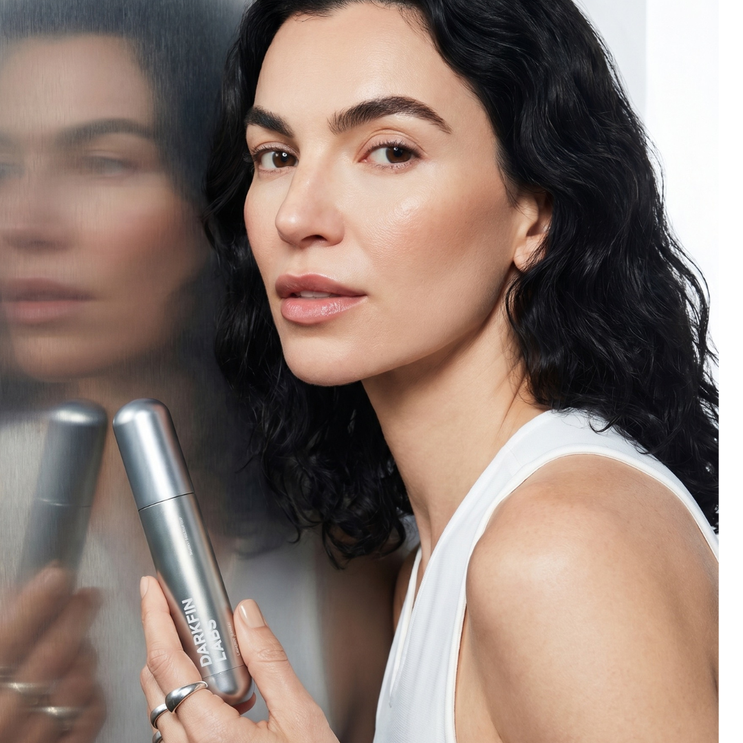 A woman with dark wavy hair holds a silver bottle, her reflection visible next to her.
