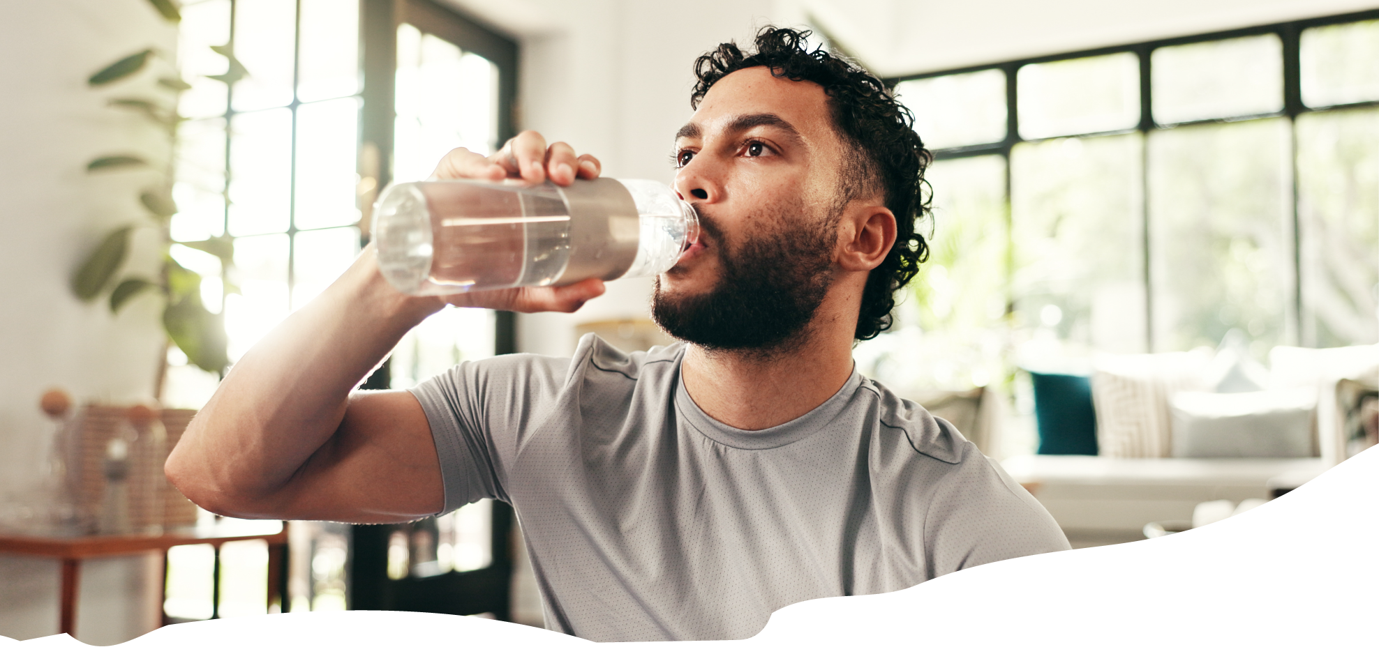 Man in a gray shirt drinking water from a bottle indoors.