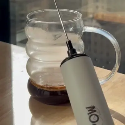 A handheld frother beside a clear glass mug with liquid on a wooden surface.