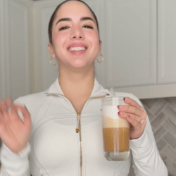 Person in a white jacket holding a glass of layered coffee in a kitchen.
