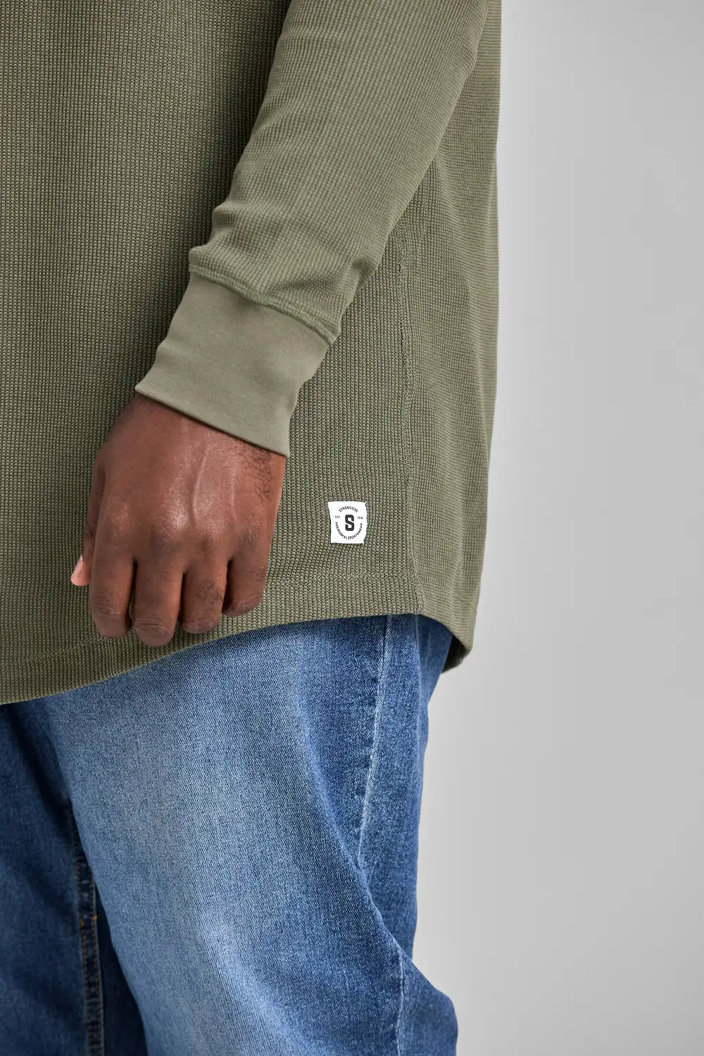 A close-up of a person wearing an olive green waffle-knit long-sleeve shirt and blue jeans.
