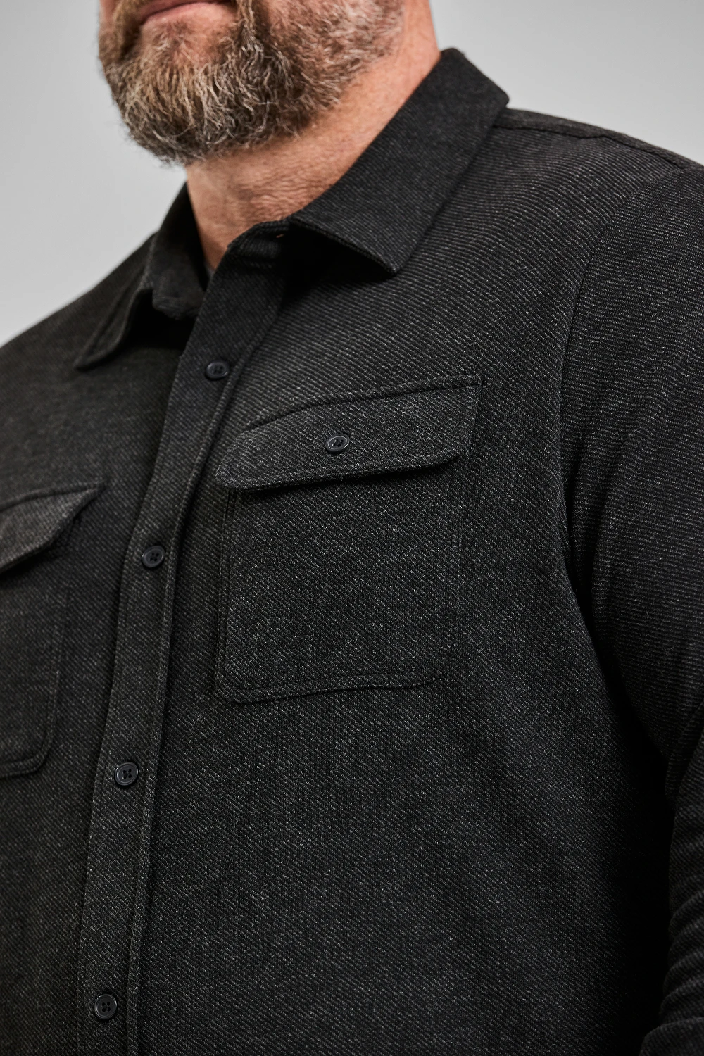 A close-up of a bearded man wearing a dark grey, textured, long-sleeved shirt with two chest pockets.
