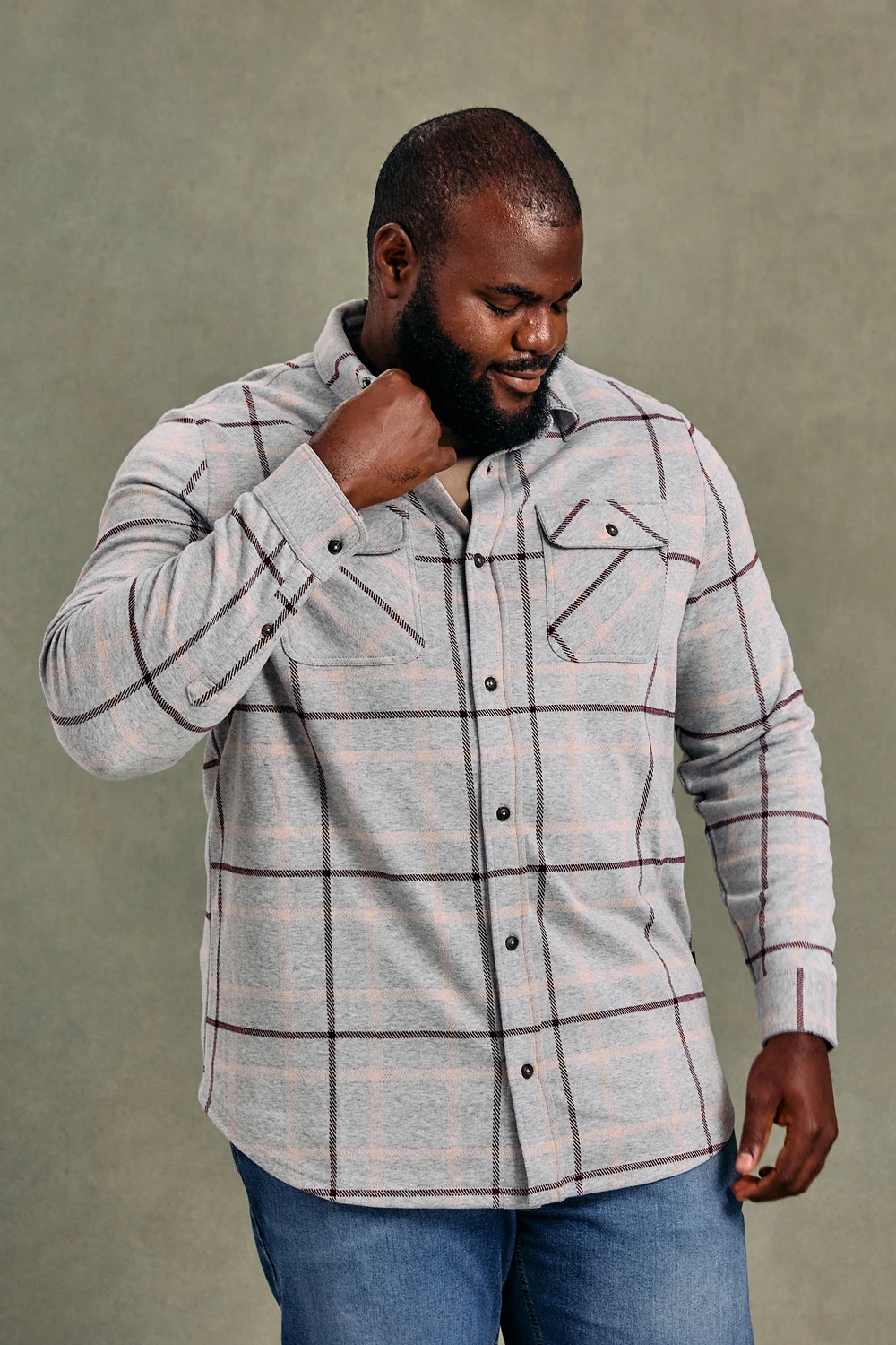 A man with a beard wearing a grey plaid button-down shirt and blue jeans looks down.