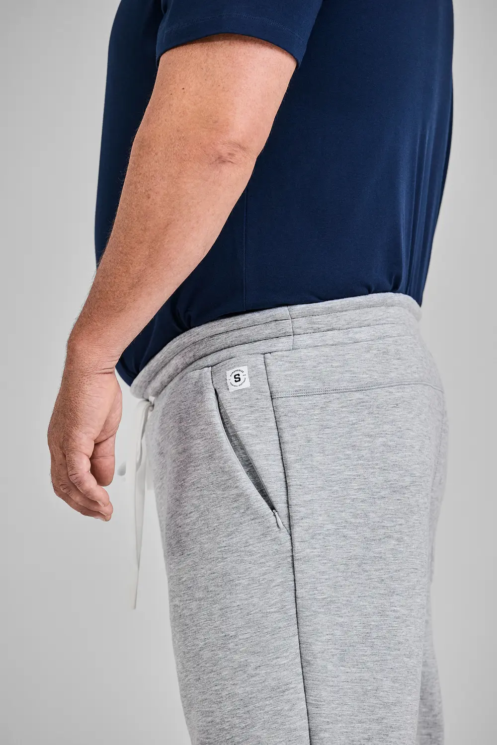 A close-up side view of a man wearing a navy blue t-shirt and grey sweatpants with zippered pockets.
