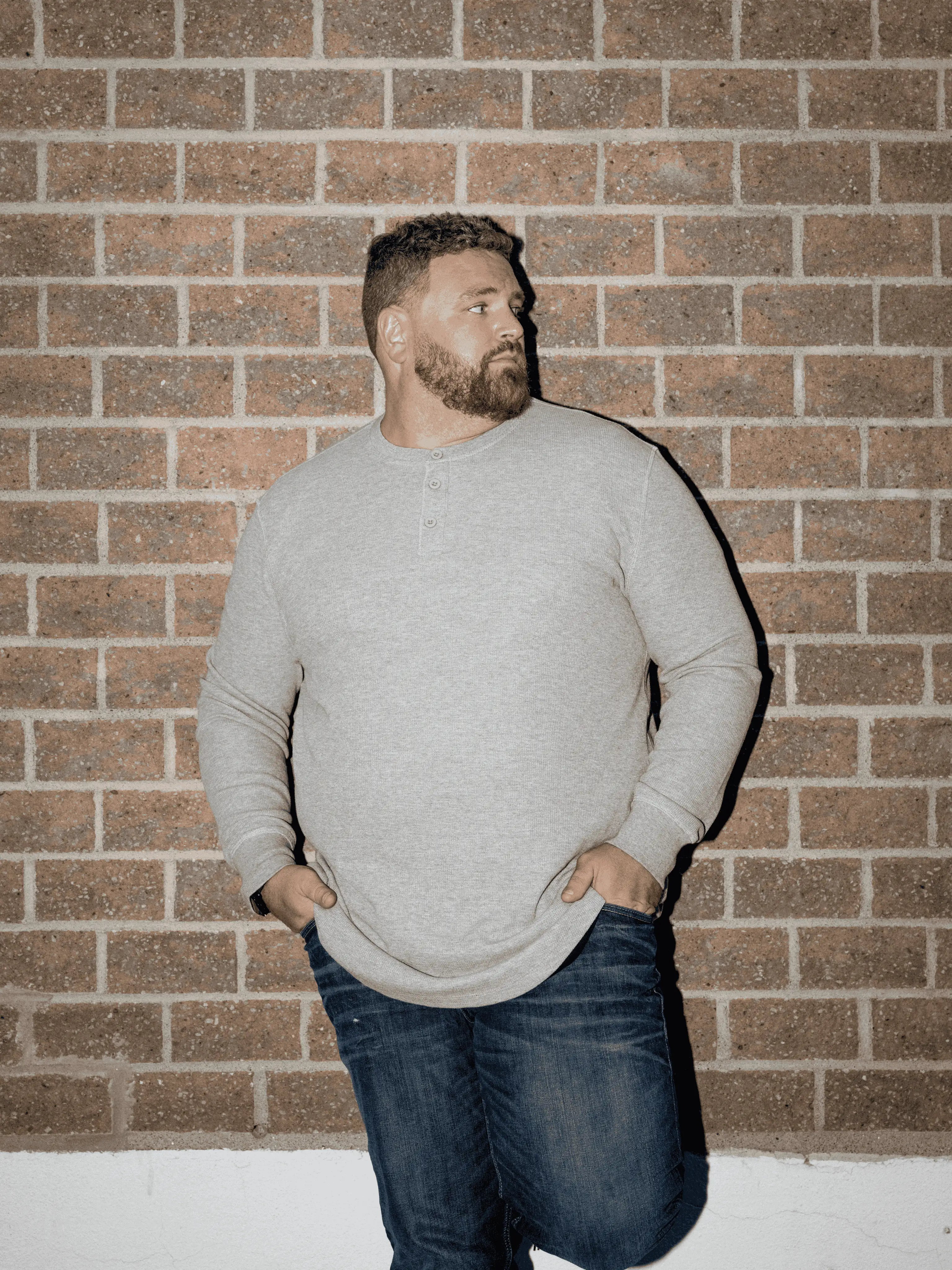 A man with a beard in a grey shirt and jeans stands against a brick wall, looking off-camera.