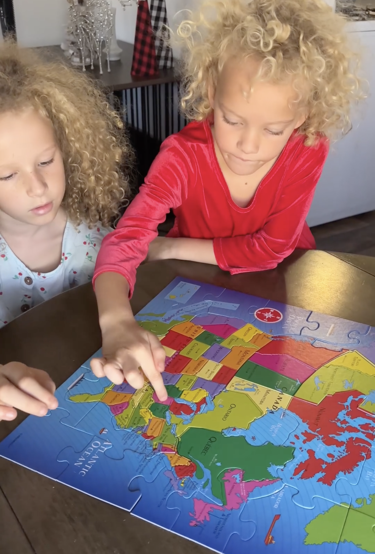 Two young girls with curly blonde hair work together on a colorful puzzle map of the United States.