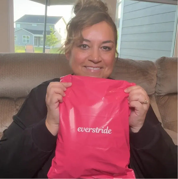 A woman sits on a couch smiling and holding up a pink Everstride mailer bag.