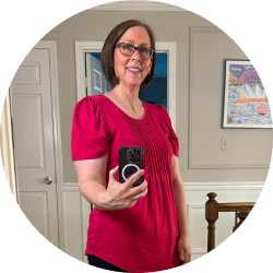 A smiling woman with glasses and short brown hair in a pink top takes a mirror selfie.