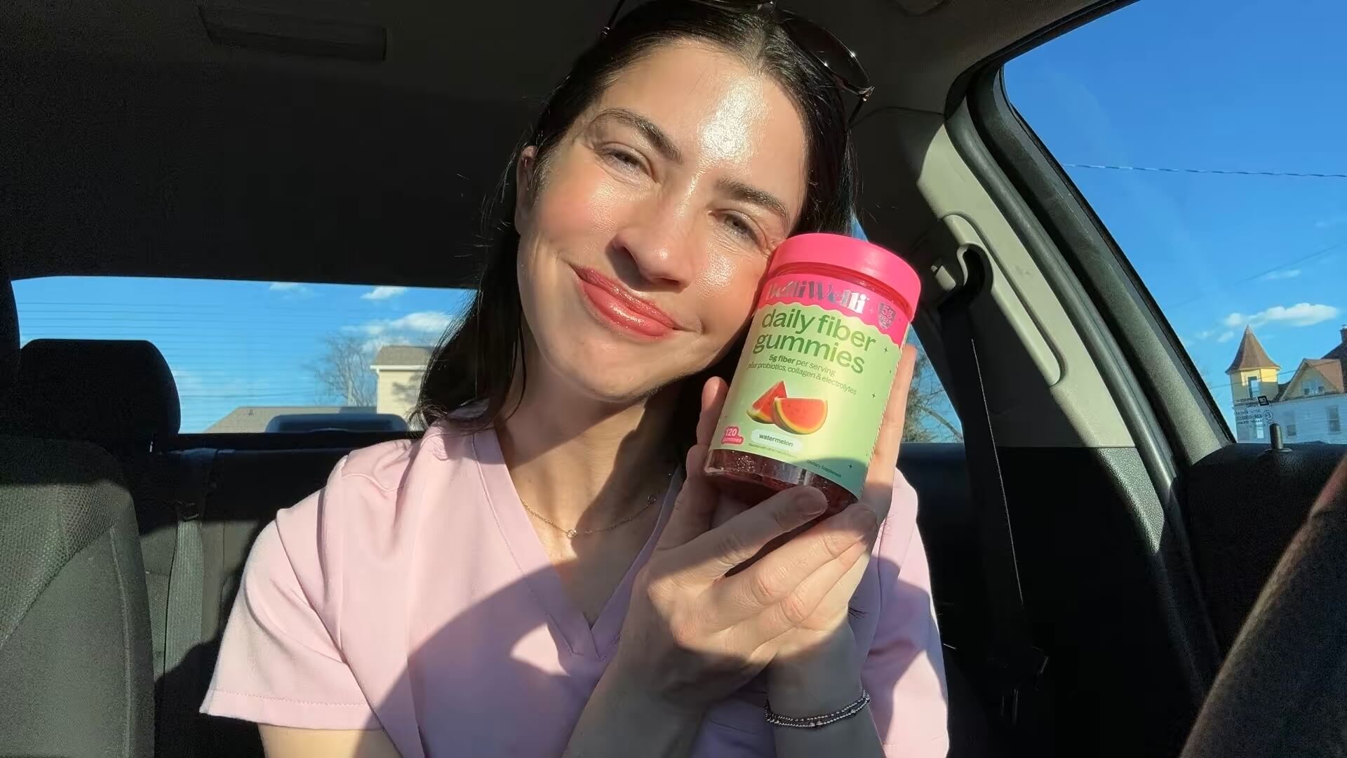 A woman sits in a car on a sunny day, smiling and holding a bottle of daily fiber gummies.