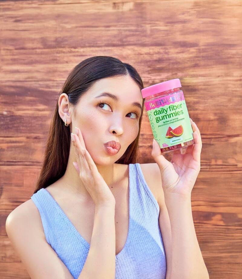A woman with a playful expression holds a jar of BelliWelli daily fiber gummies against a wood background.