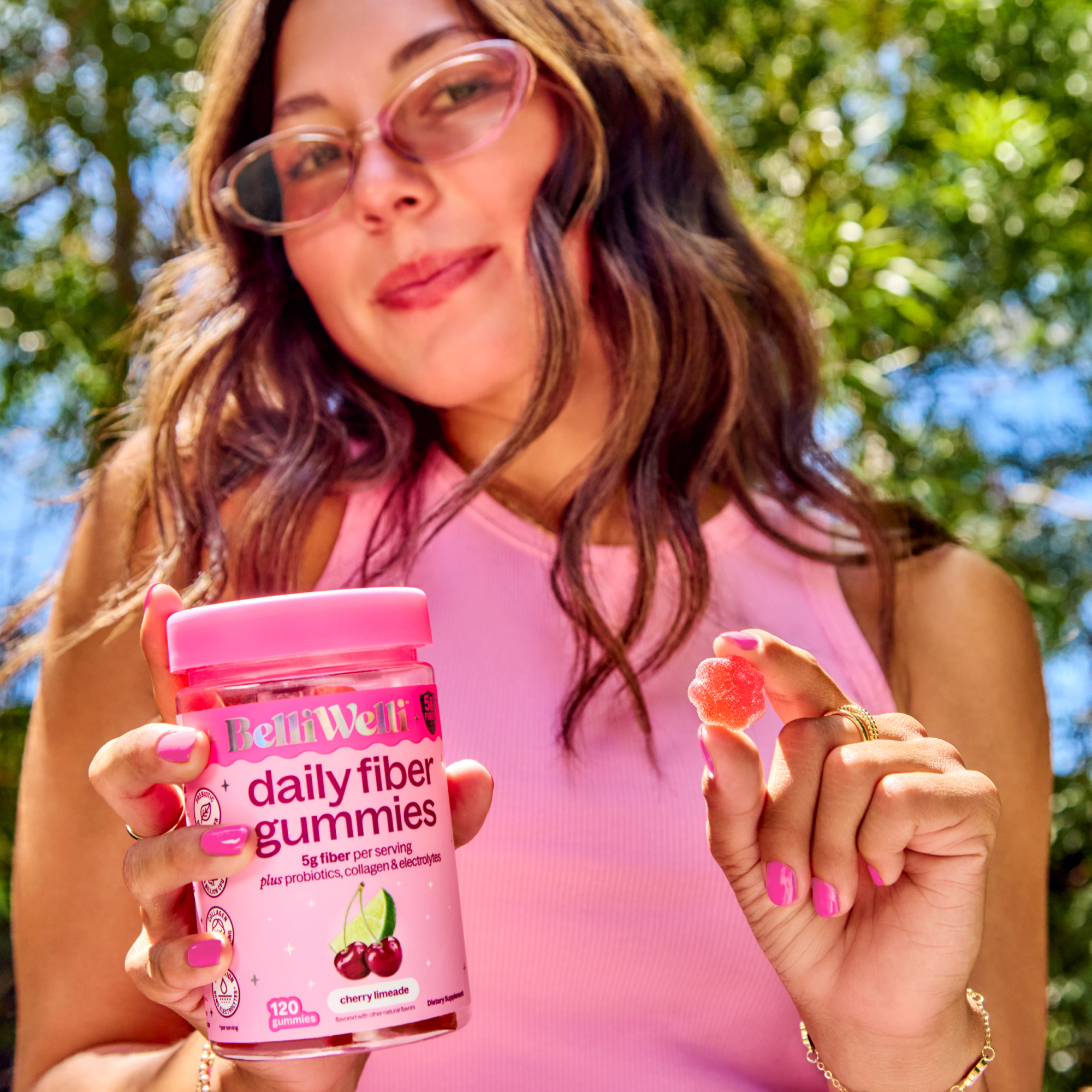 A woman holds a bottle of BelliWelli daily fiber gummies and a single red gummy.