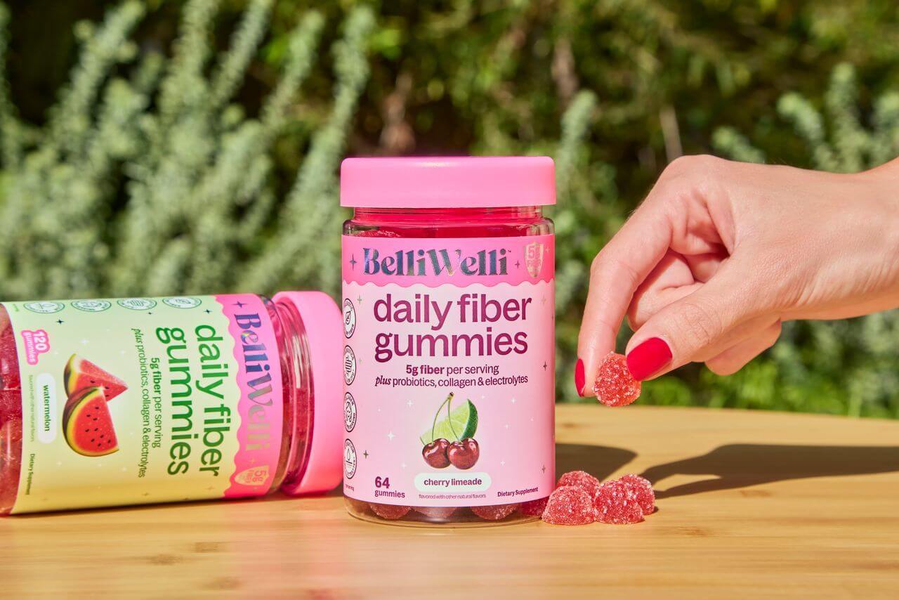 A hand with red nail polish picks up a BelliWelli daily fiber gummy from a wooden table.
