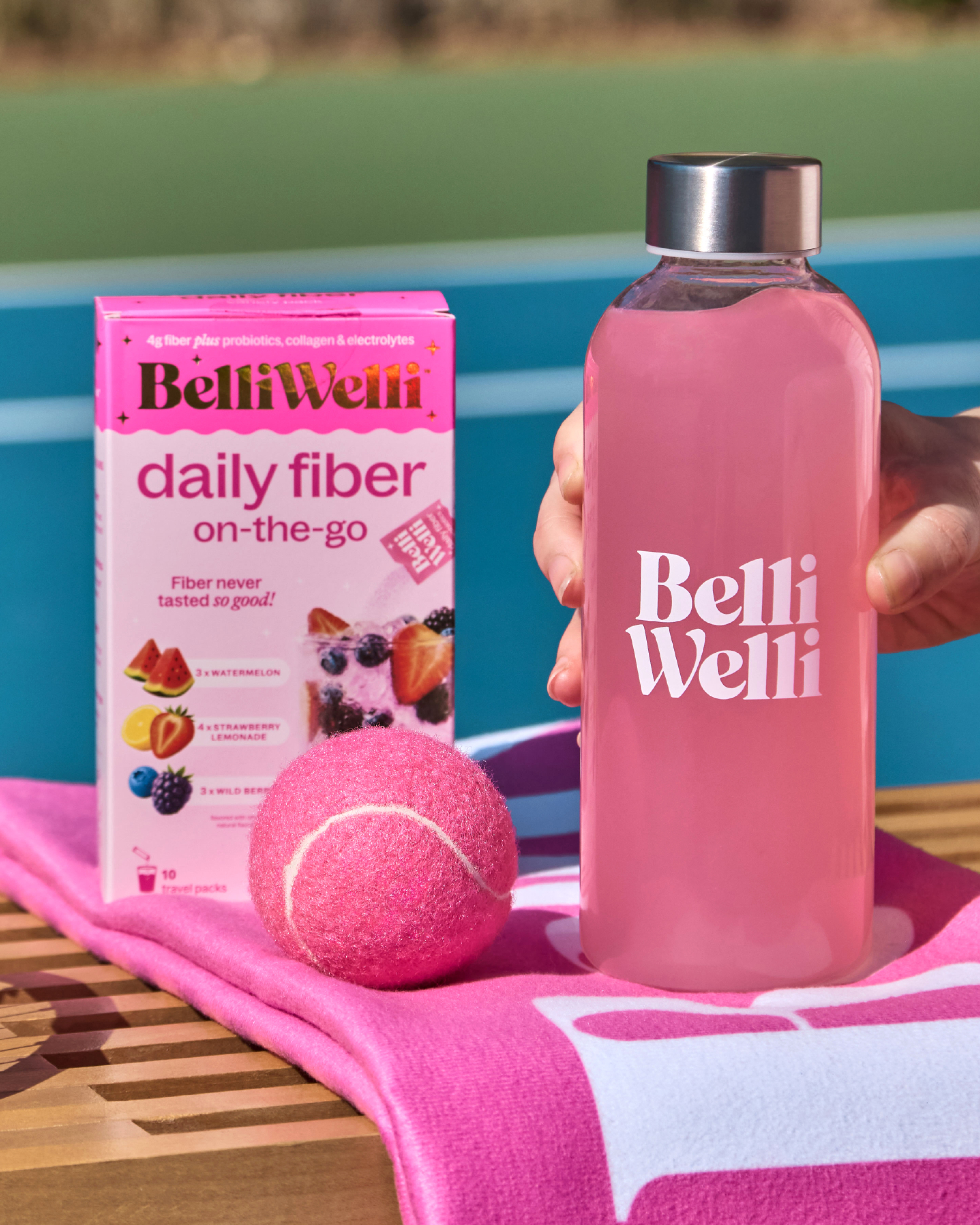 A box of BelliWelli daily fiber, a bottle of pink drink, and a pink tennis ball on a towel.