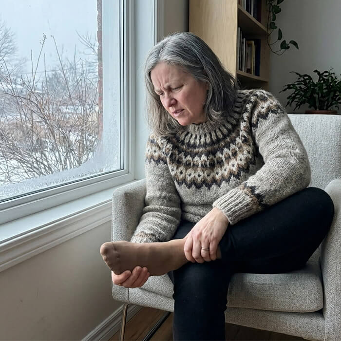 A woman in a sweater sits in a chair, holding her stockinged foot with a pained expression.