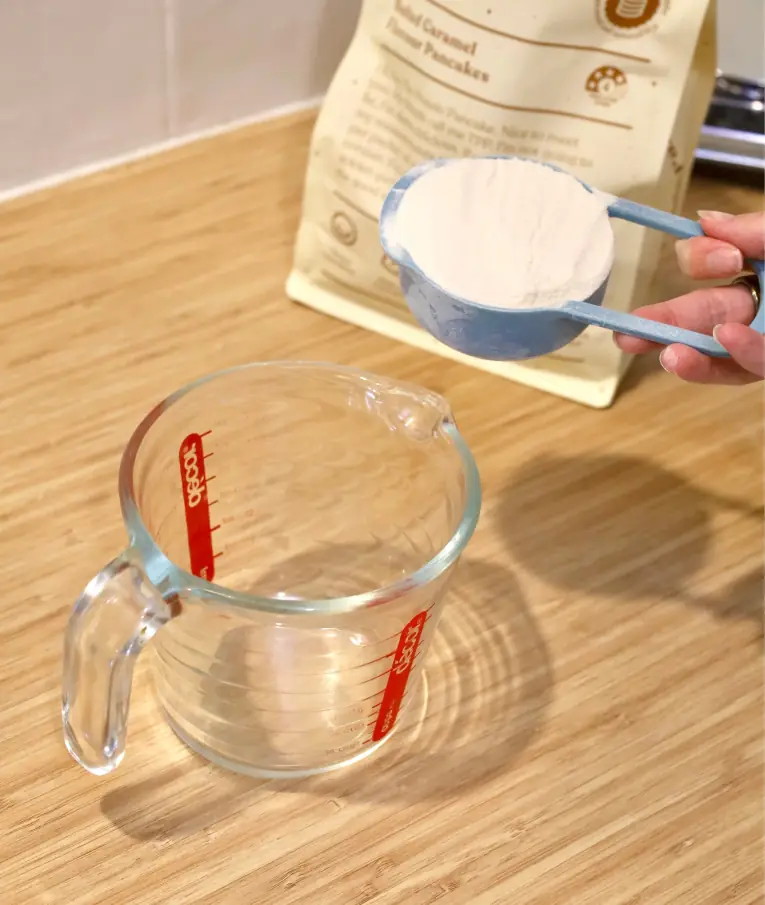 Person holding measuring cup with flour above empty glass jug on wooden countertop.