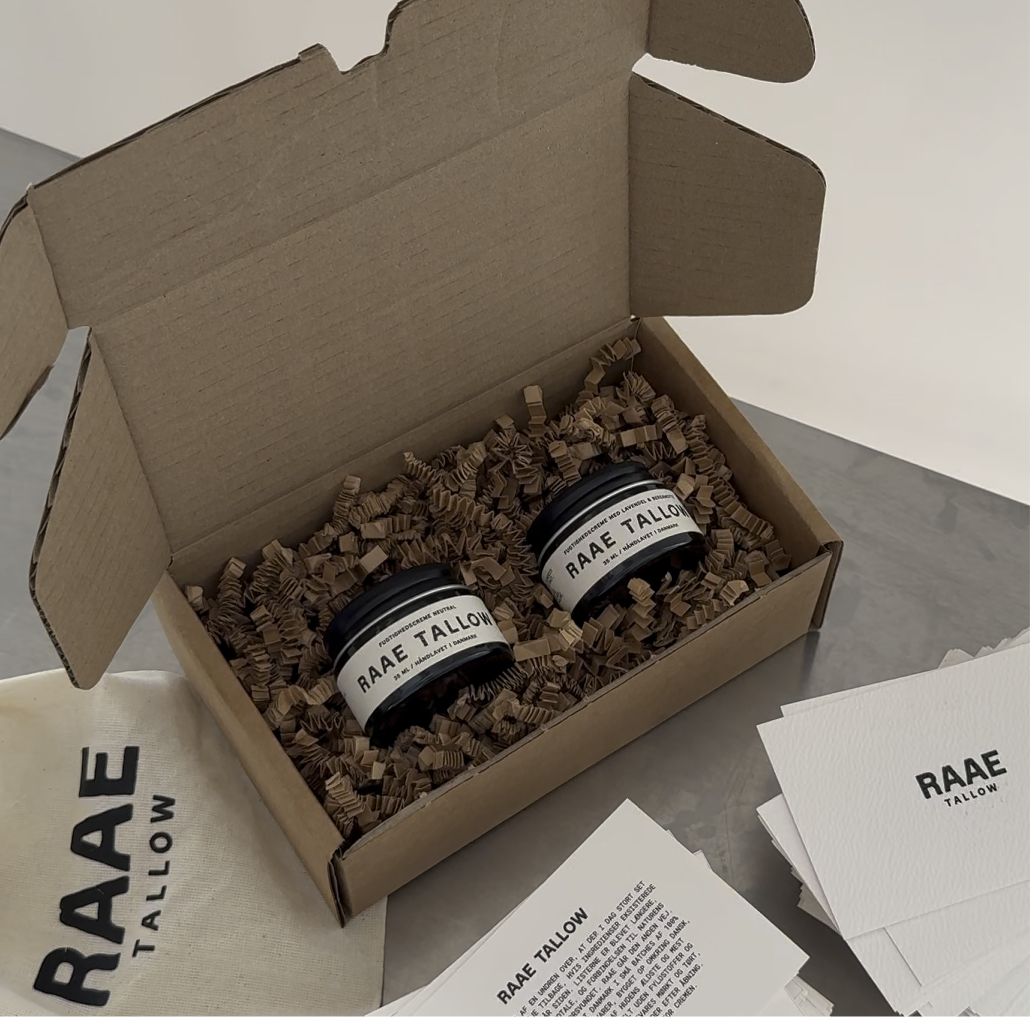 An open box containing two jars labeled 'RAAE TALLOW' surrounded by brown paper shred.