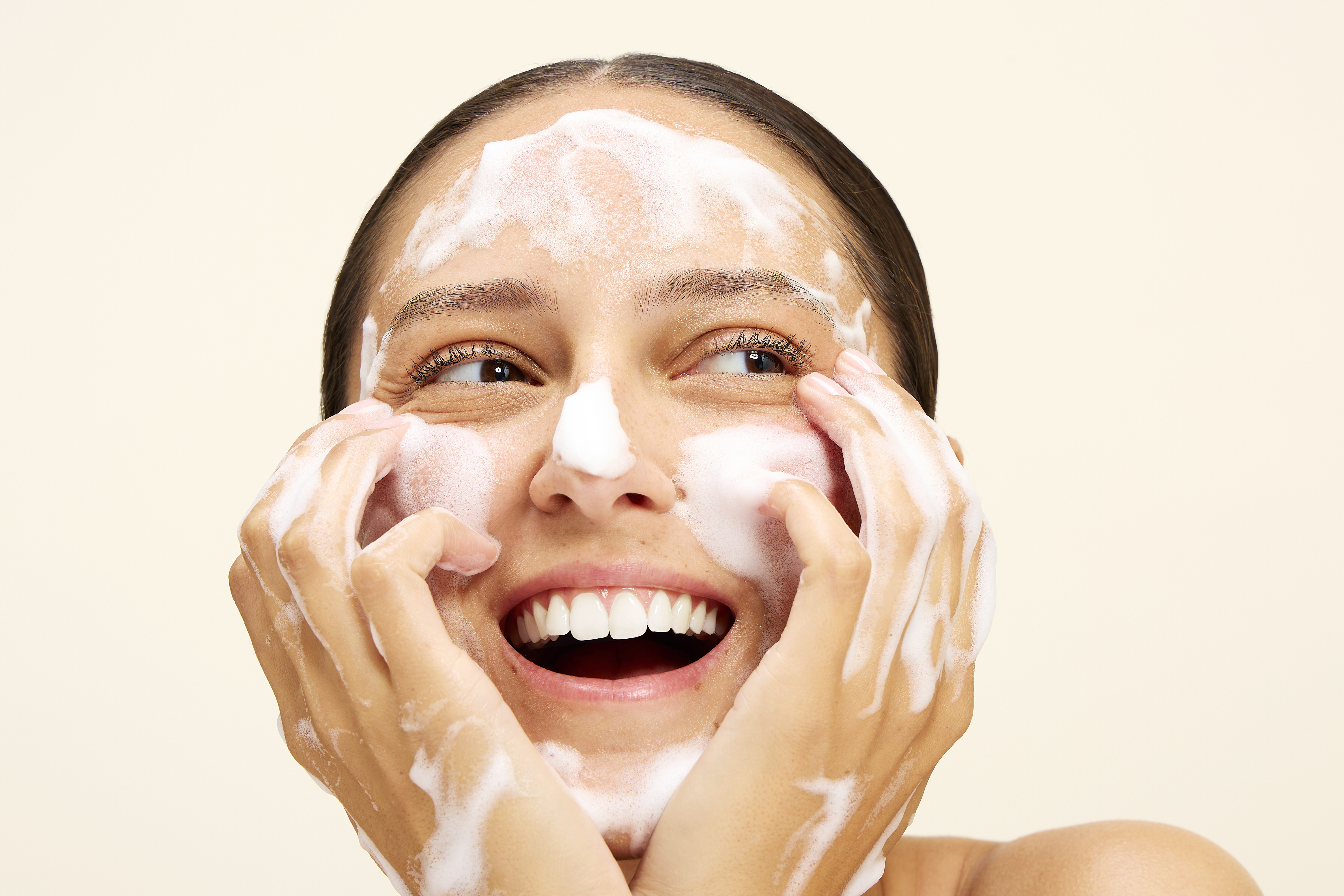A smiling woman washes her face with a foaming cleanser against a cream background.