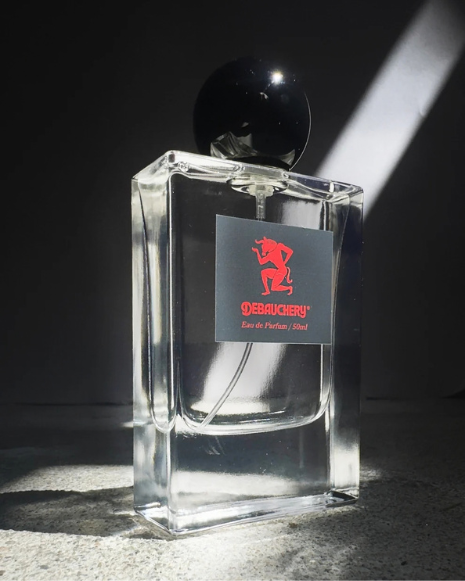A rectangular glass bottle of Debauchery perfume with a black cap and a red devil logo.