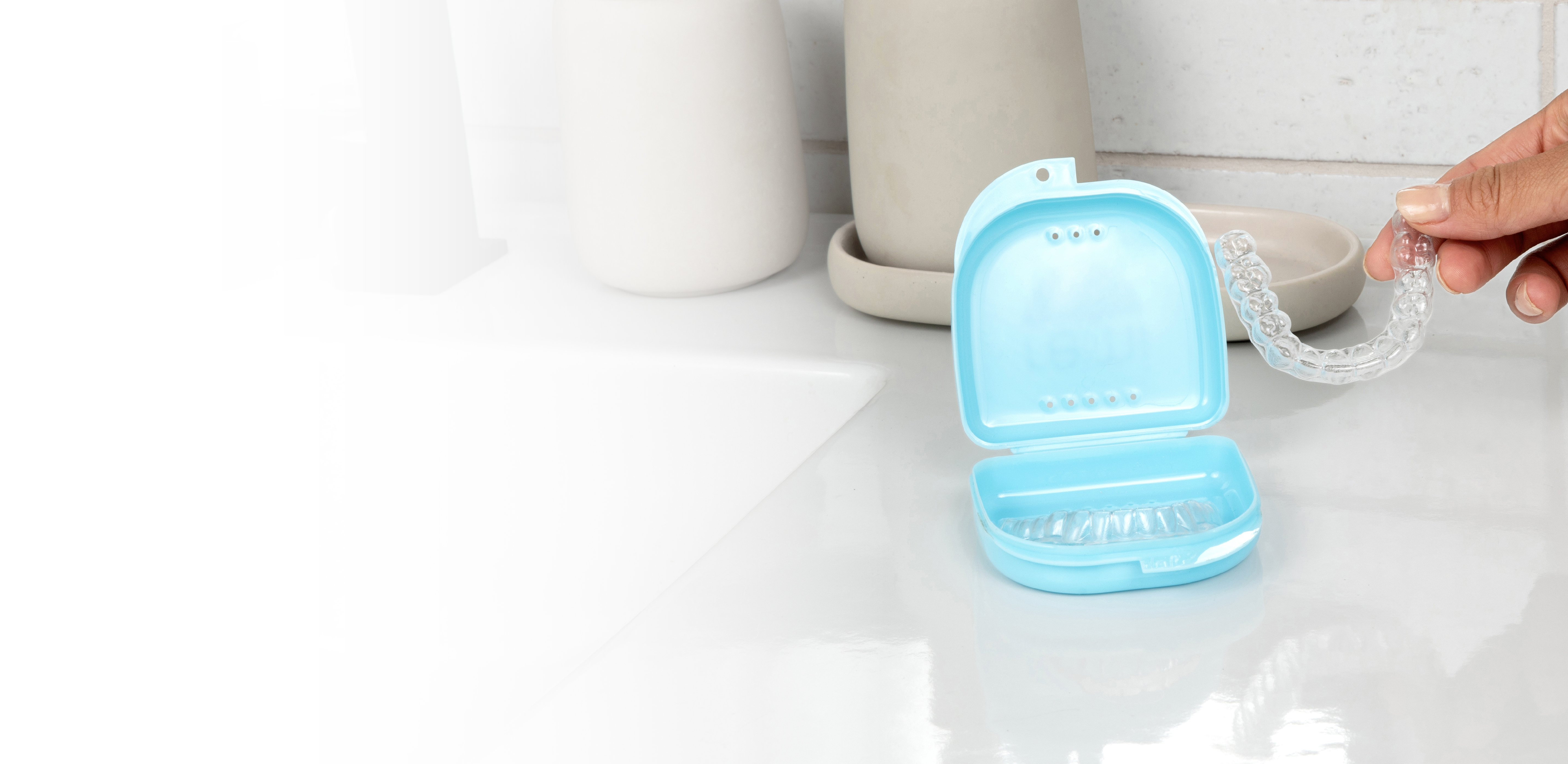 Hand holding a clear dental aligner above a blue case on a bathroom counter.