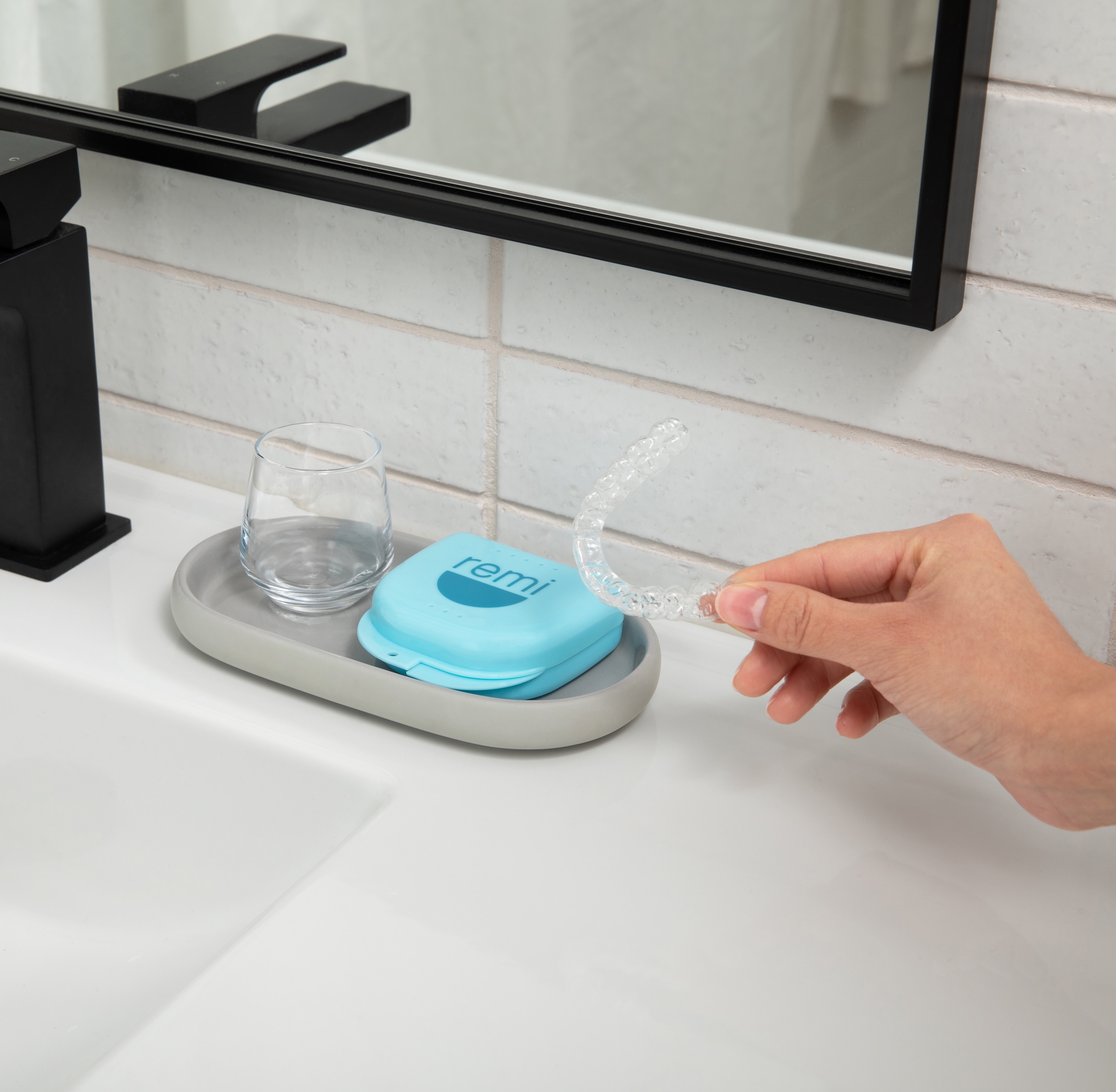 Hand holding a clear dental retainer near a sink with a case and glass.