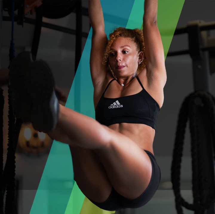 A woman in a black Adidas sports bra and shorts performing a hanging leg raise in a gym.
