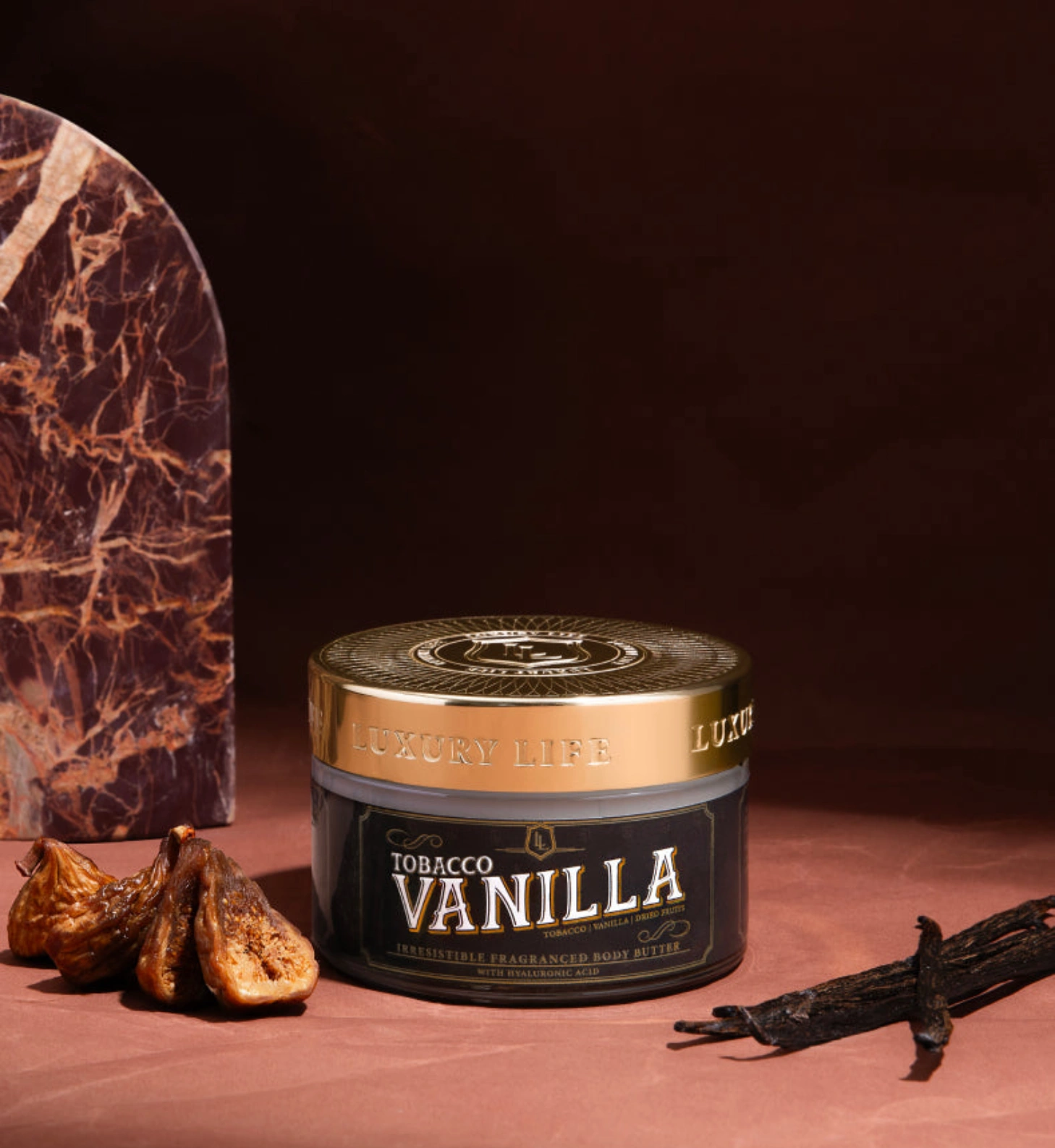A jar of Tobacco Vanilla body butter next to dried figs and vanilla beans on a dark background.