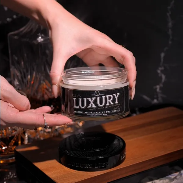 A pair of hands holds an open glass jar of Luxury brand fragranced body butter.