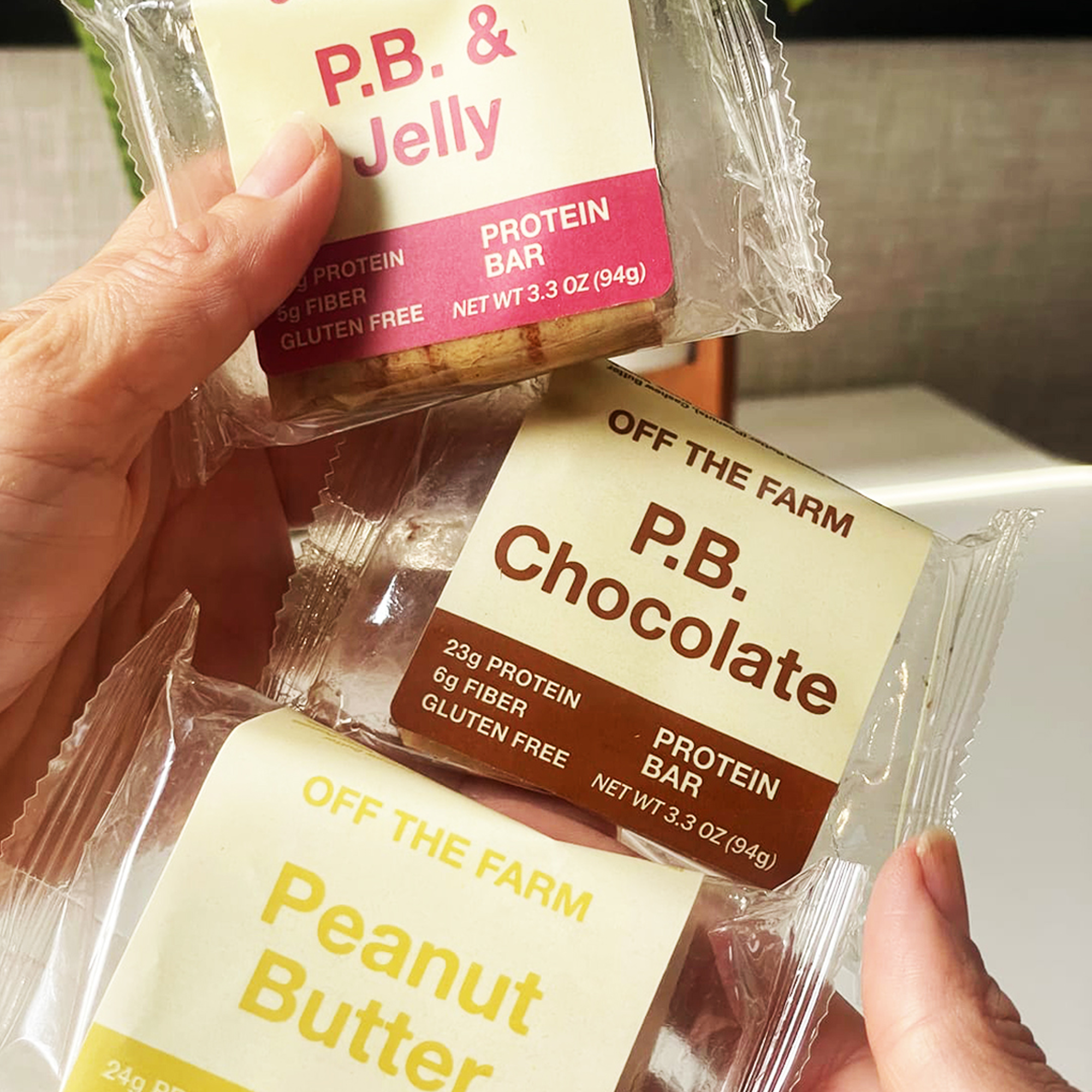 Hands holding three different protein bars labeled P.B. & Jelly, P.B. Chocolate, and Peanut Butter.