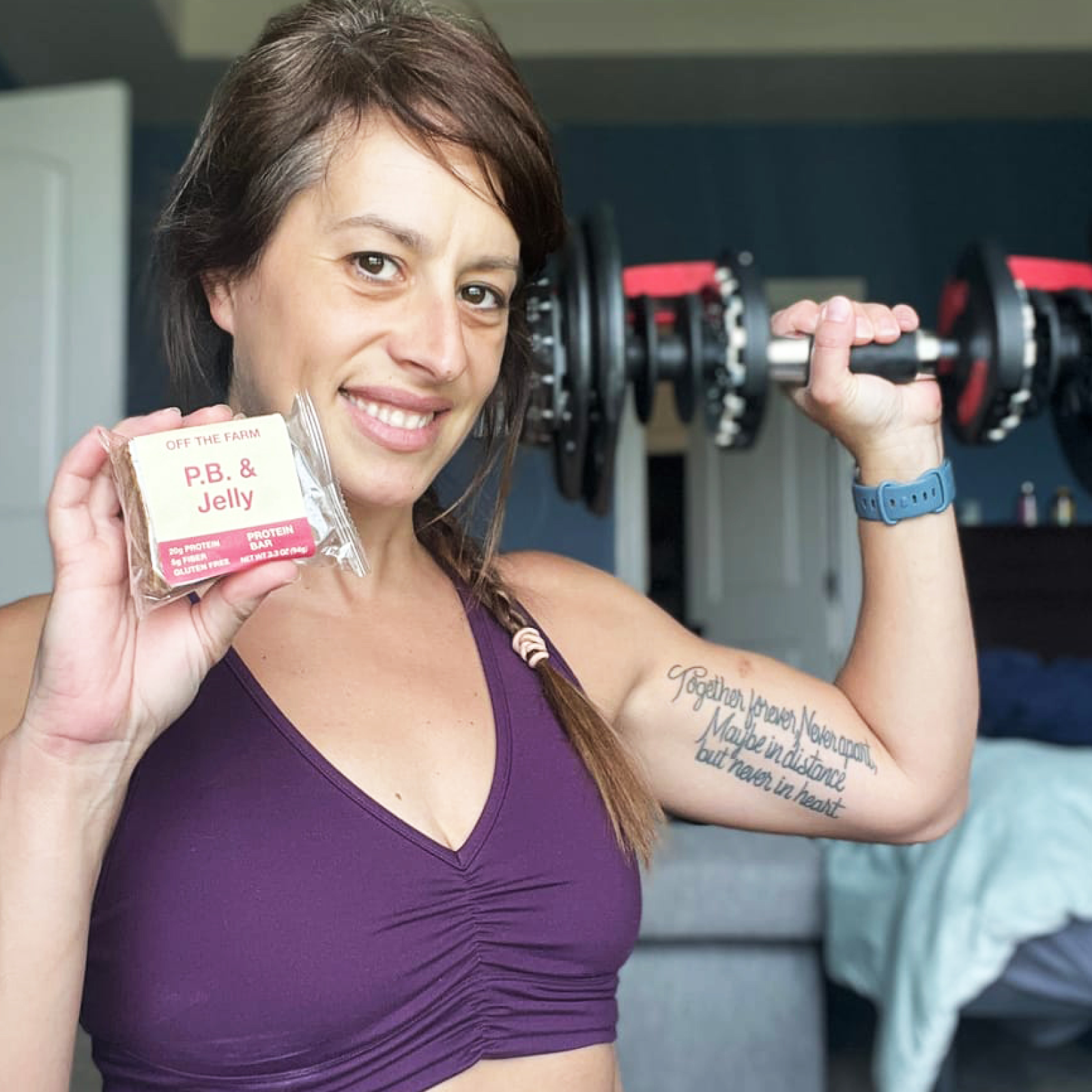 Woman holding a protein bar and a dumbbell, smiling in a workout setting.