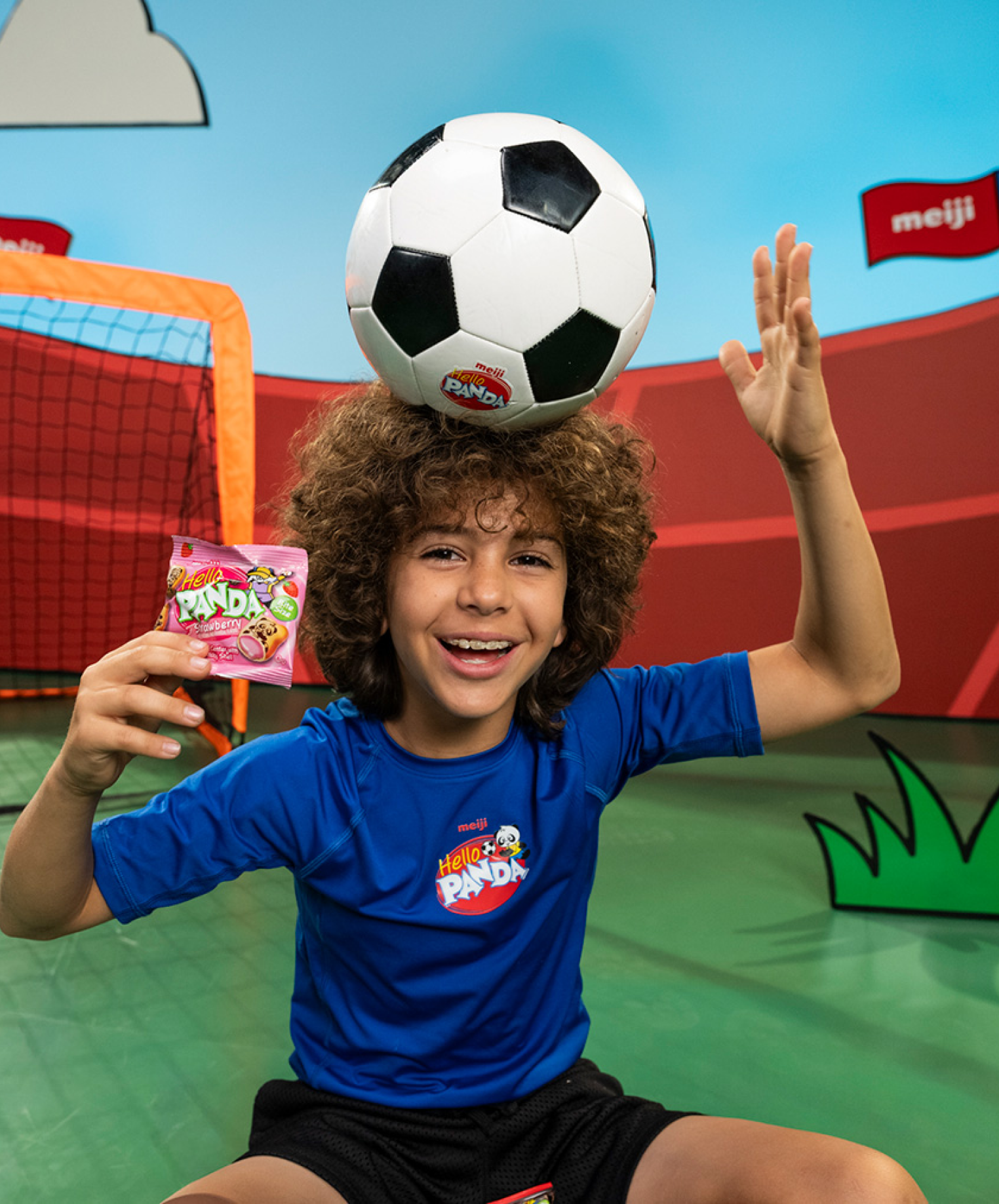 Child balancing soccer ball on head, smiling, holding snack pack.