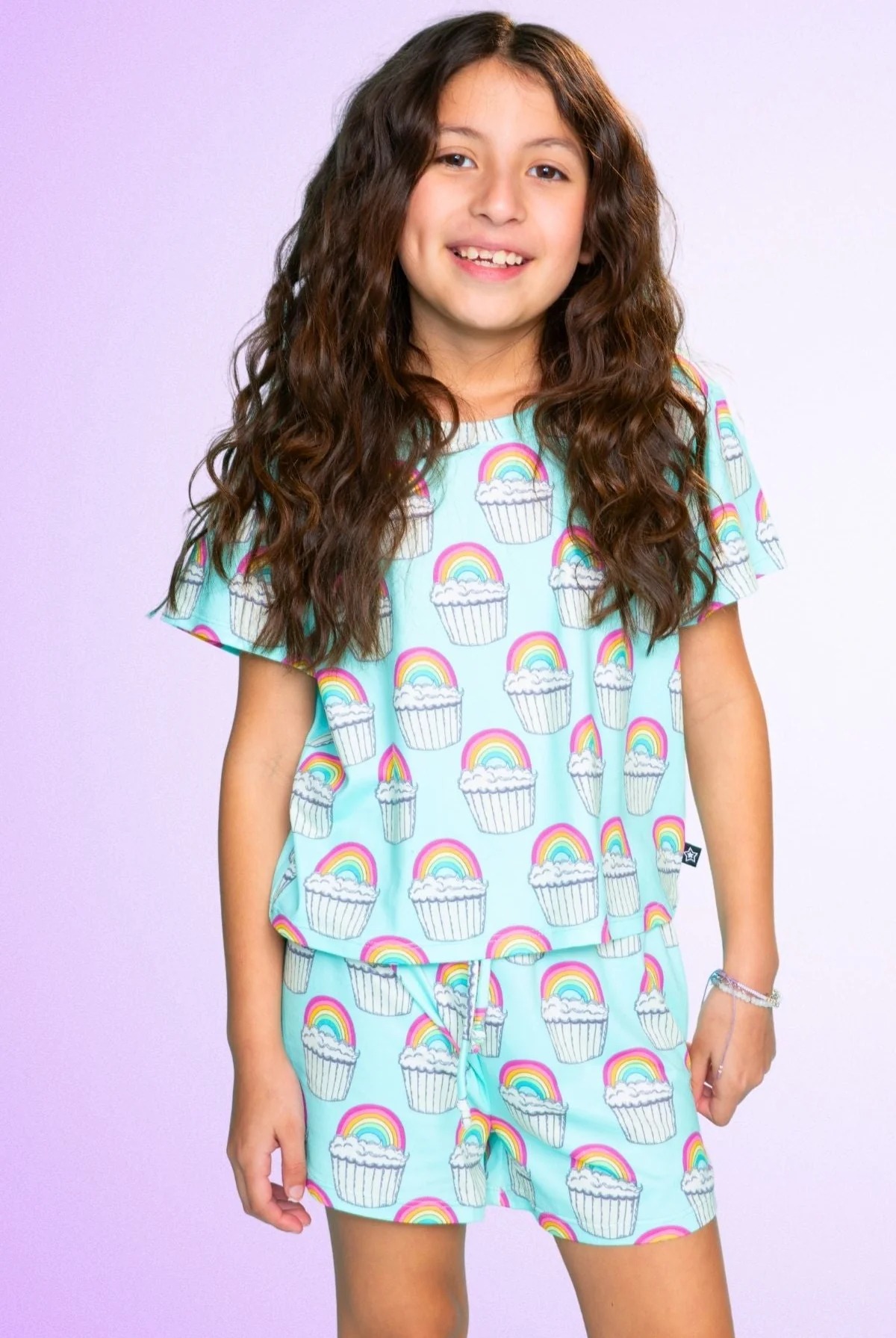 Child wearing rainbow cupcake patterned outfit on a purple background.