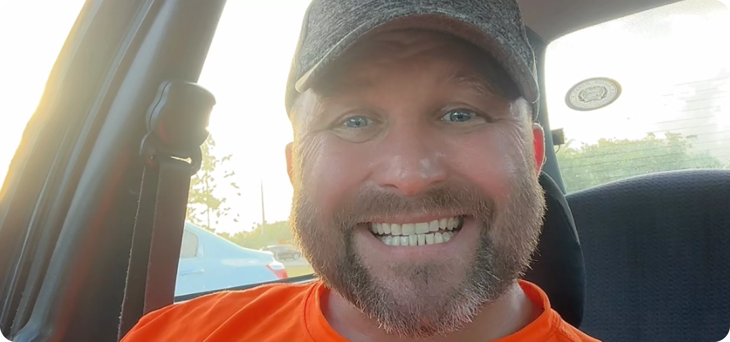 Smiling man in a car wearing an orange shirt and a cap.