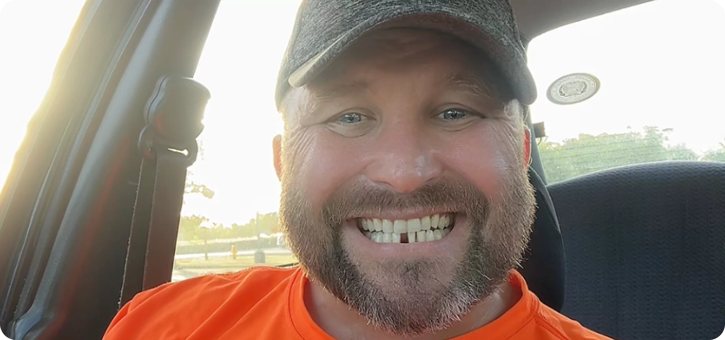 Smiling man in a car wearing an orange shirt and a cap.